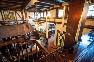 Escada de madeira com corrimão, velas sobre suporte e vista para um bar com teto de madeira e janelas.