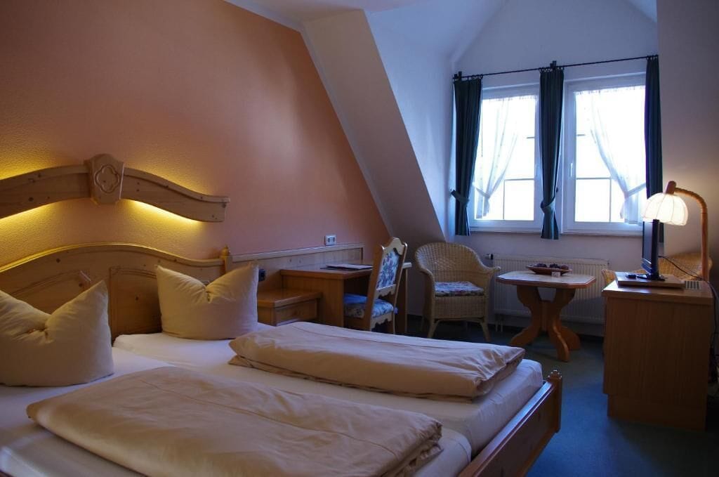 A cozy bedroom with a sloped ceiling, a bed with white linens, a chair, a small table, and a lamp.