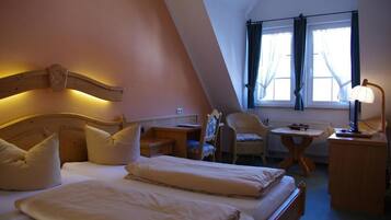 A cozy bedroom with a sloped ceiling, a bed with white linens, a chair, a small table, and a lamp.