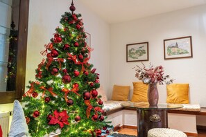 Ein Weihnachtsbaum mit roten Verzierungen und Lichtern, ein Glastisch, eine Vase mit Blumen und ein Sofa mit gelben Kissen.