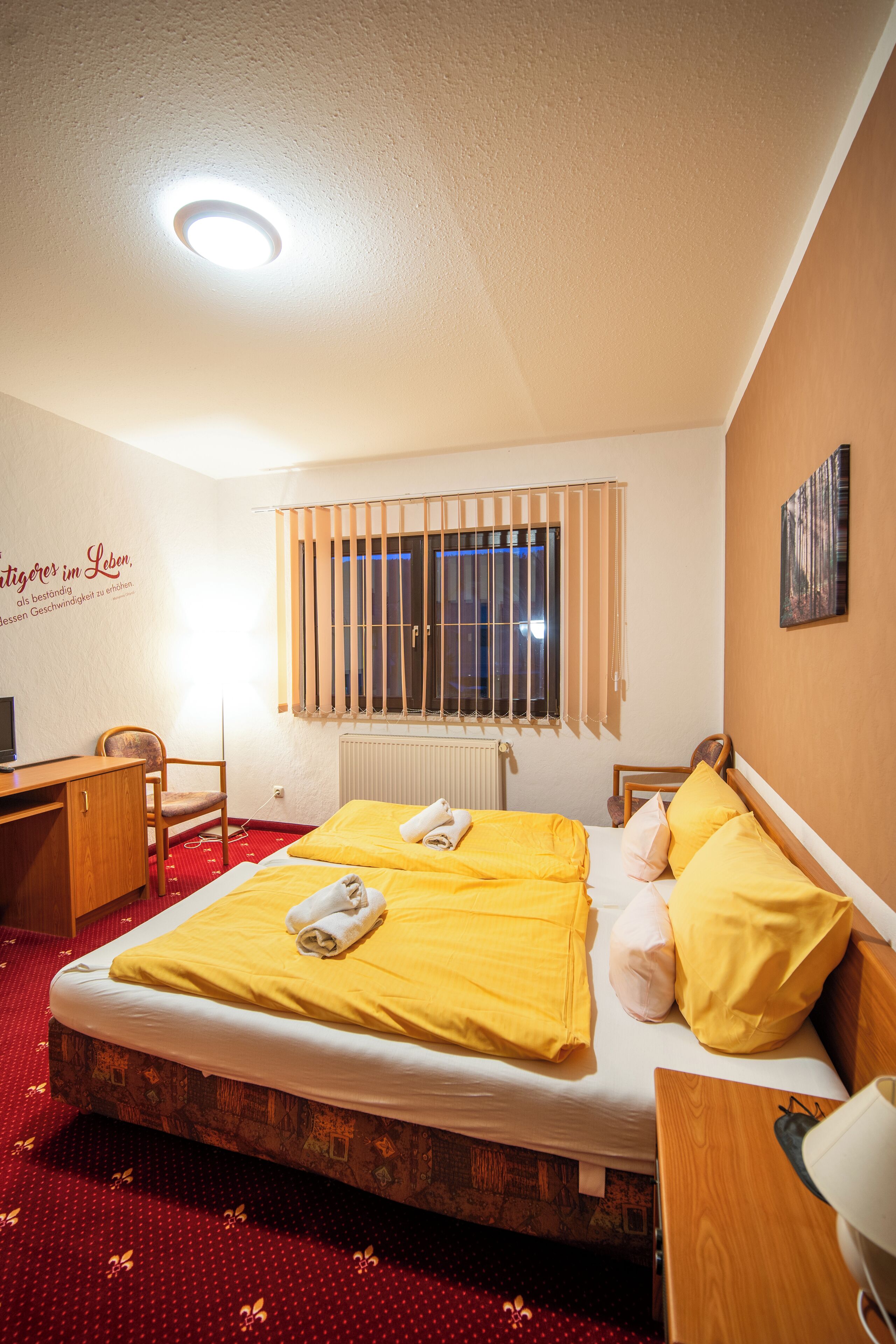 A hotel room with a bed, a desk, a chair, a radiator, and a window with bars.