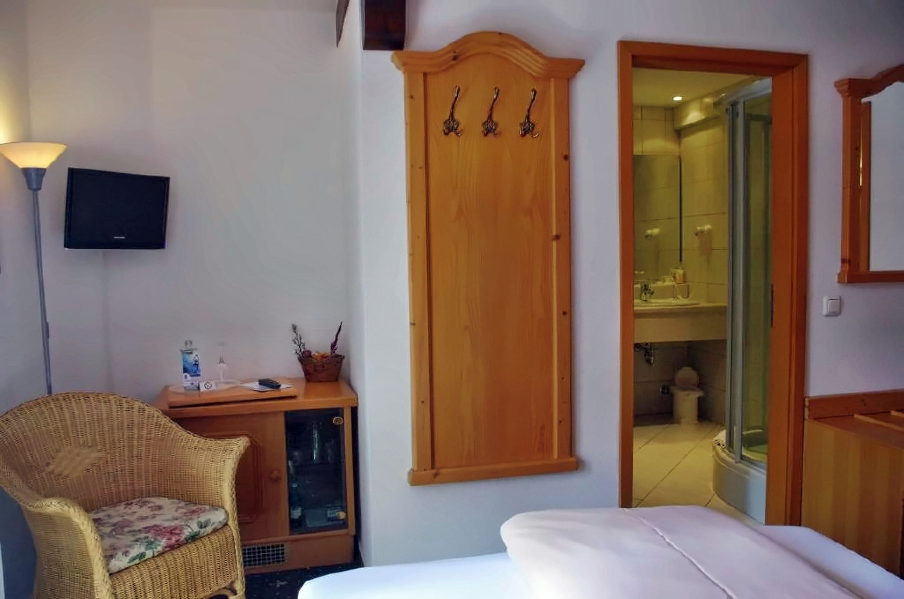 Una habitación con cama, una silla de mimbre, un armario de madera, televisión y un baño a la vista a través de una puerta abierta.