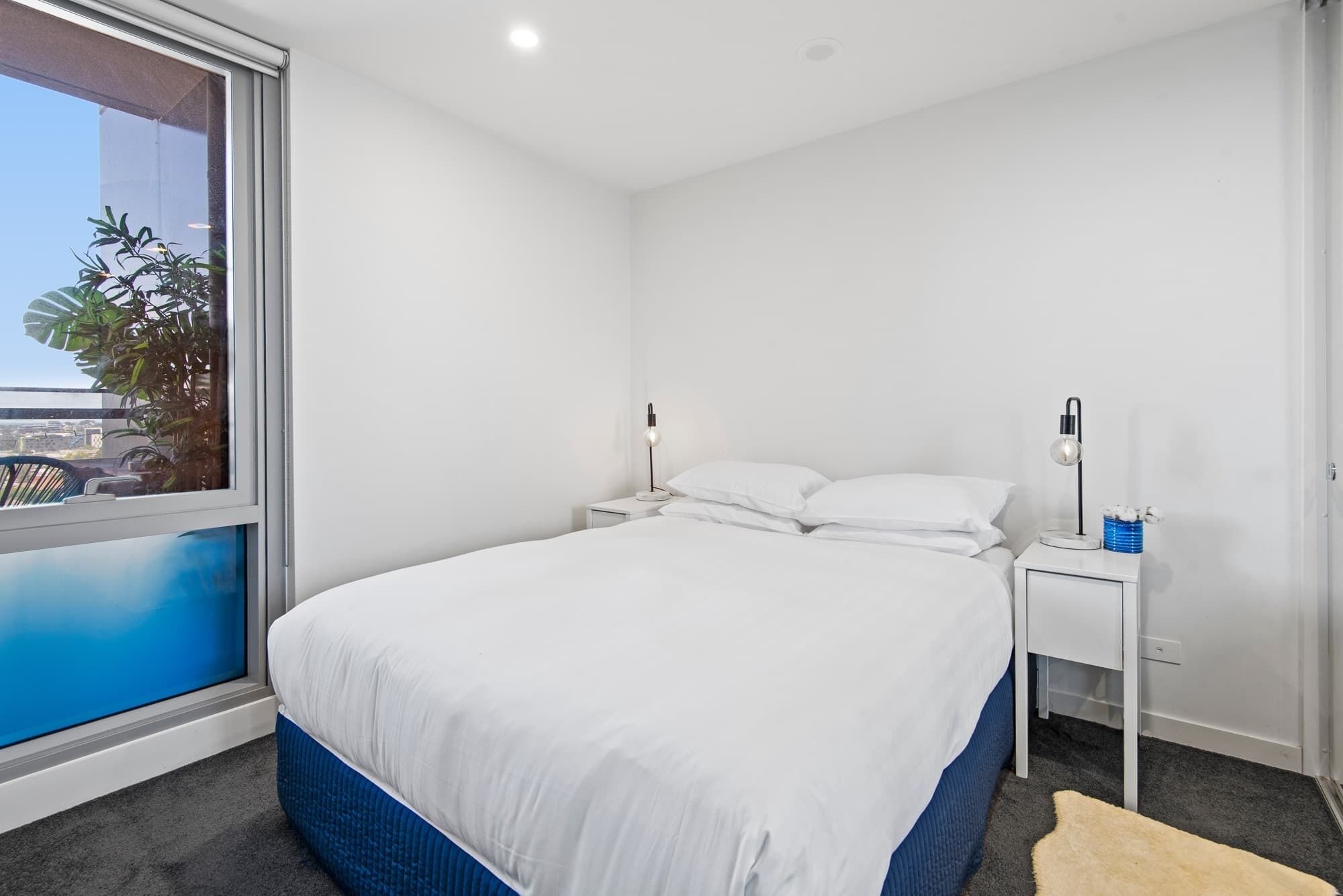 A bedroom with a large bed, white walls, and a window with a view of buildings and greenery.
