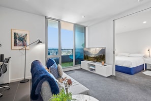 Enjoy a city view from the balcony through the large sliding glass door. The room features a comfortable seating area, flat-screen TV, and a neatly made bed.
