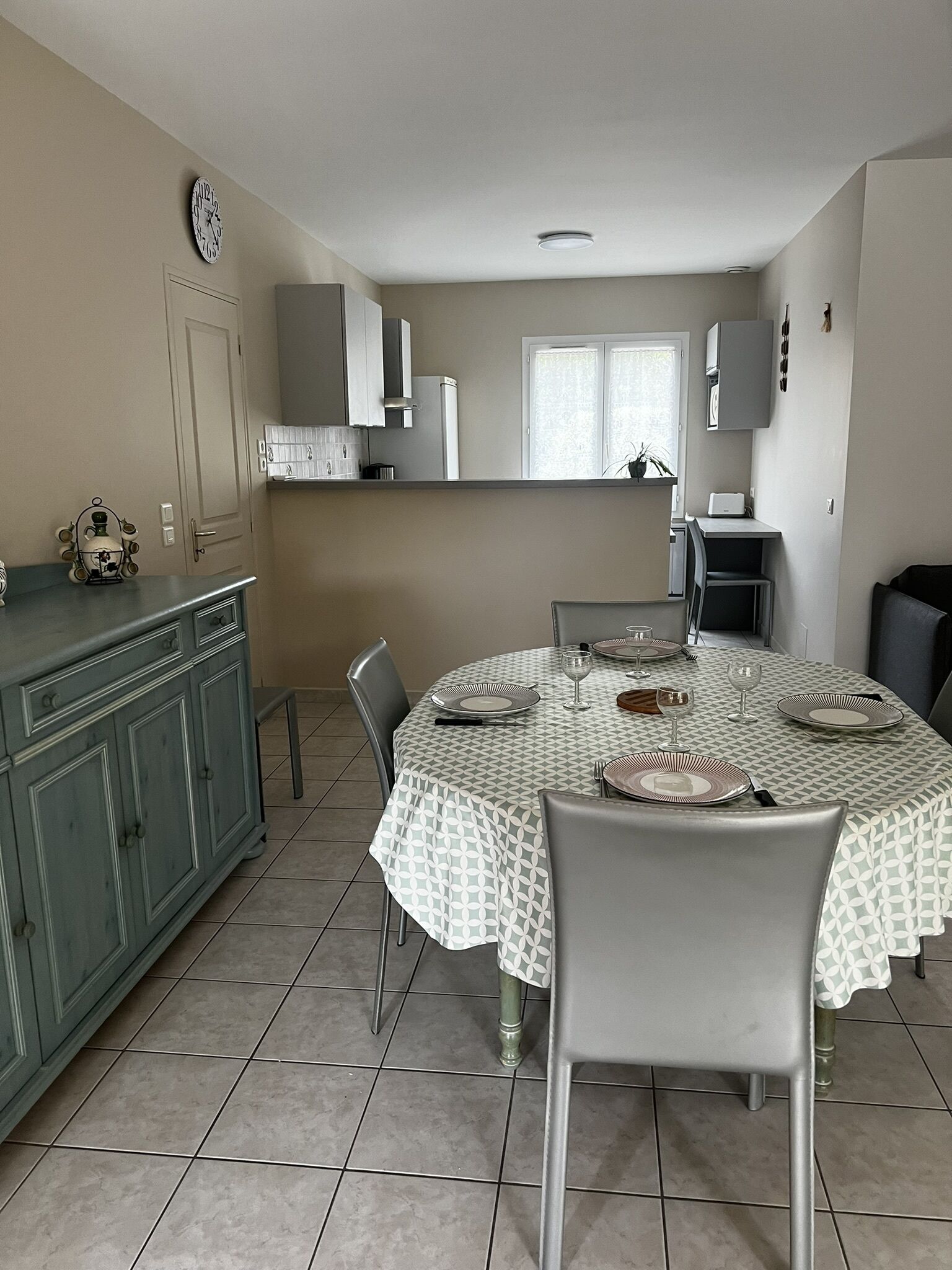 A dining area with a round table set for four, a kitchen in the background, and a clock on the wall.