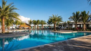 A swimming pool with palm trees and lounge chairs, surrounded by buildings with a tropical theme.