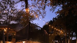 Un chemin de pierre mène à une terrasse en bois dotée d’une rambarde, le tout étant entouré d’un espace paysager agrémenté d’arbres et de guirlandes lumineuses.