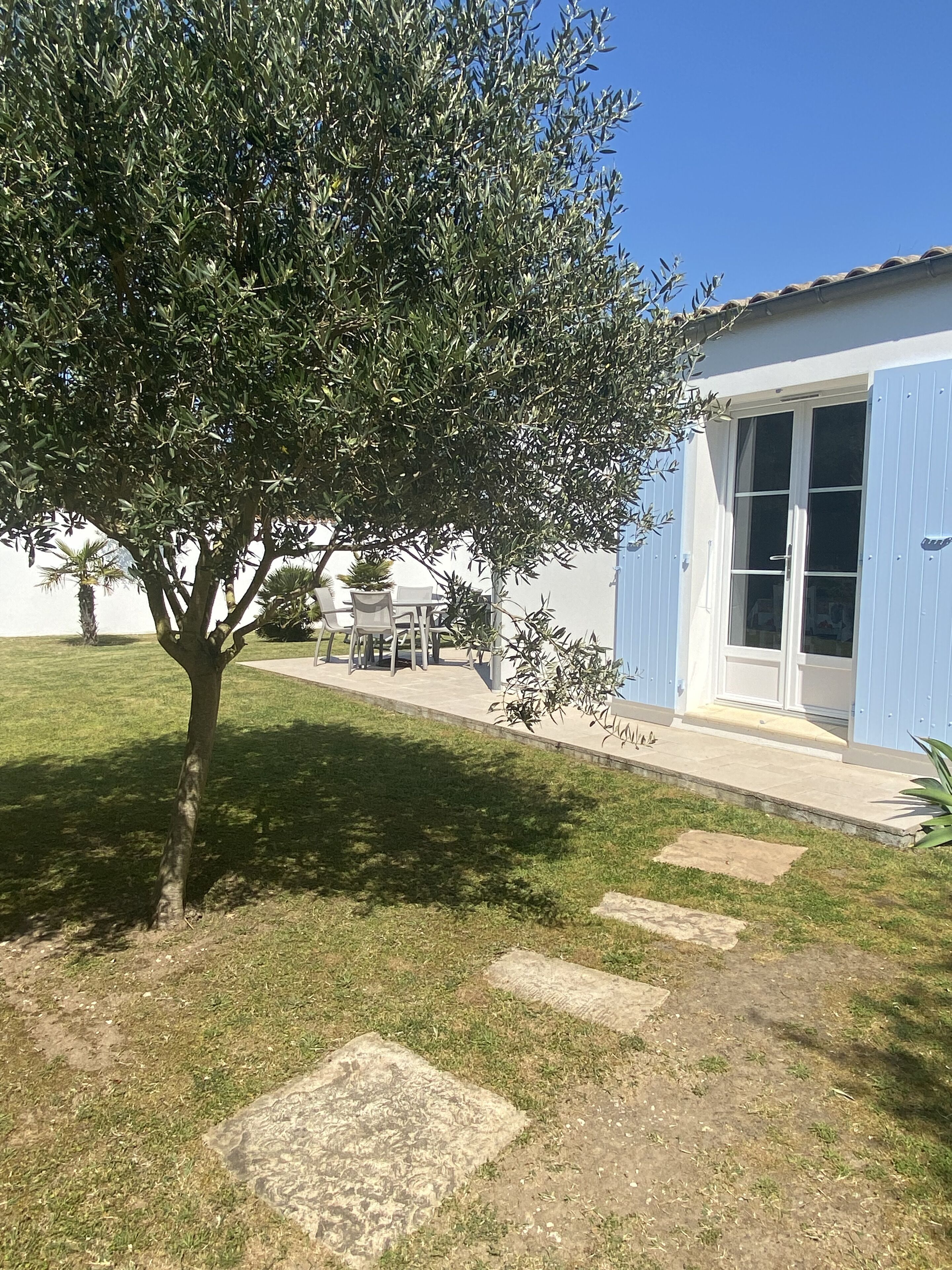 Ein Haus mit blauer Fassade, ein kleiner Garten mit Baum und eine Terrasse mit Gartenmöbeln.