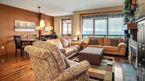A spacious living room with a fireplace, a dining area with a wooden table and chairs, and large windows providing natural light. - Ski in/out! 4 bdrms,Blacksmith Lodge,views,heated garage,internet,8 pers hot tub (Beaverdell)