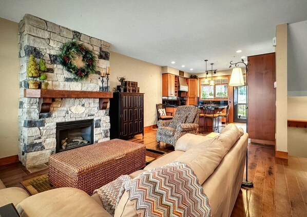 A cozy living room with a stone fireplace, a sofa set, and a dining area with a kitchen in the background. - Ski in/out! 4 bdrms,Blacksmith Lodge,views,heated garage,internet,8 pers hot tub (Beaverdell)