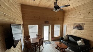 Interior de una cabaña de madera con una mesa de comedor, sillas, un sofá negro y un ventilador de techo.
