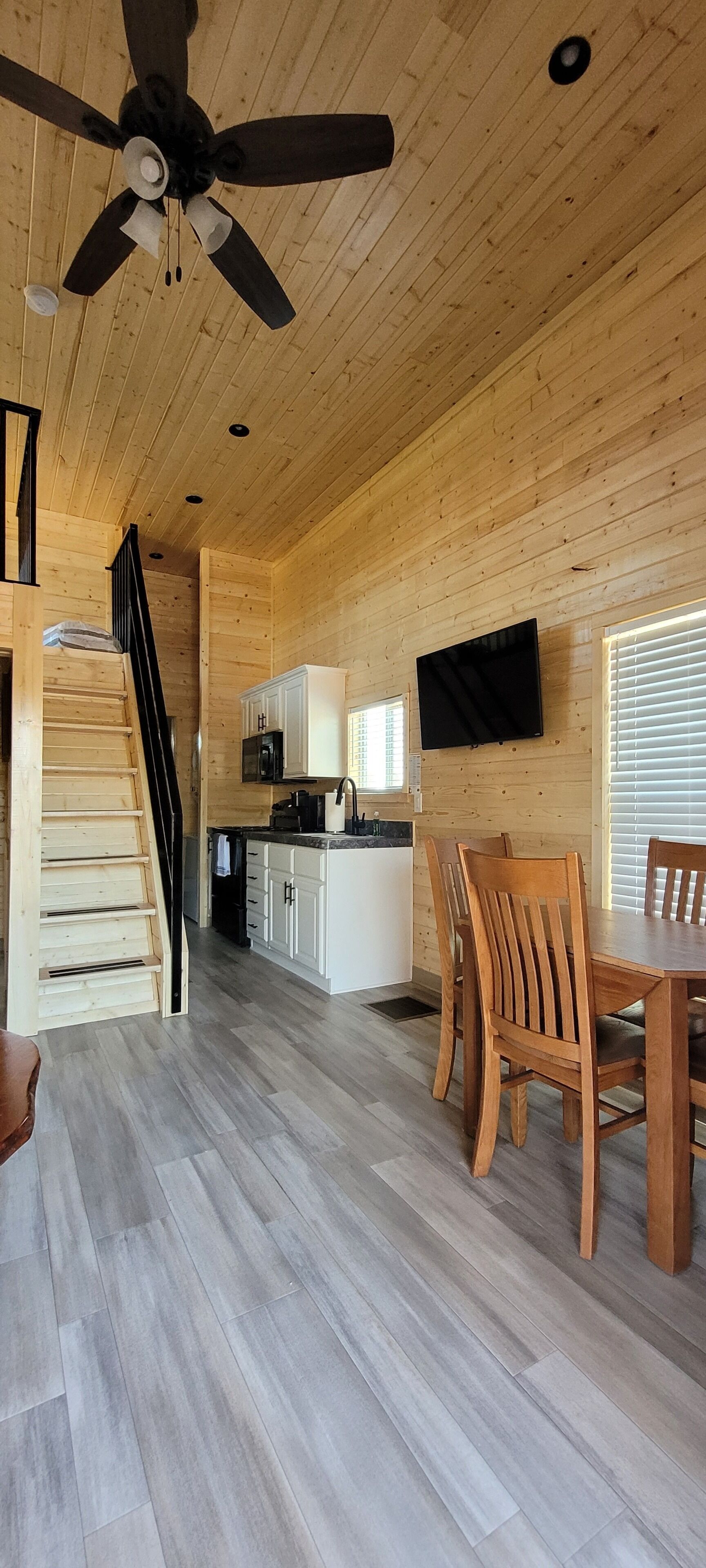 Un intérieur en bois avec un escalier, un coin repas avec des chaises en bois, une cuisine avec des meubles blancs et un ventilateur de plafond.