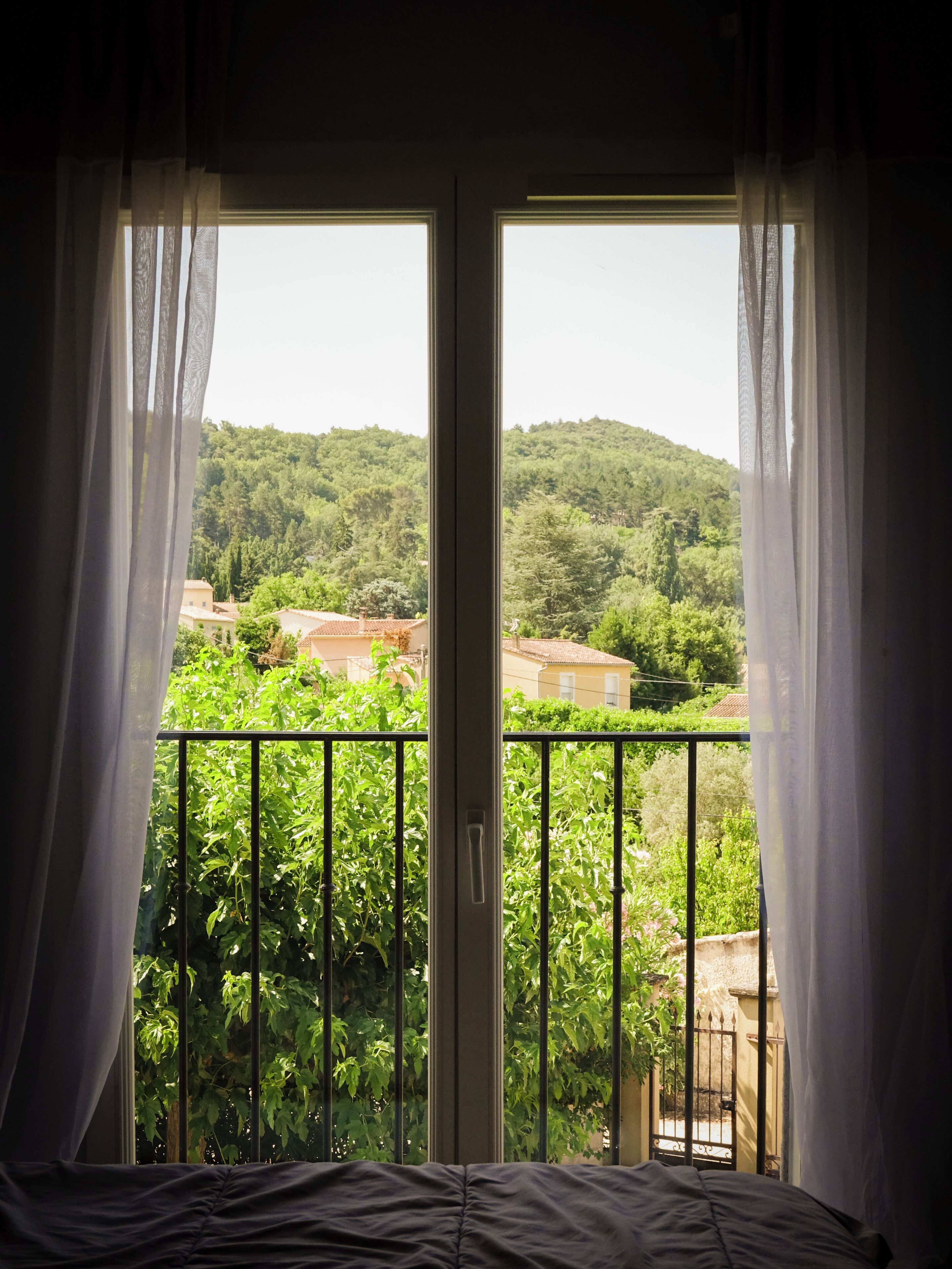 A room with a balcony, a bed, and a view of a green landscape with buildings.