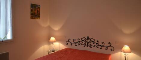 A bedroom with a bed, two bedside lamps, a decorative wall headboard, a painting, and a window with curtains.