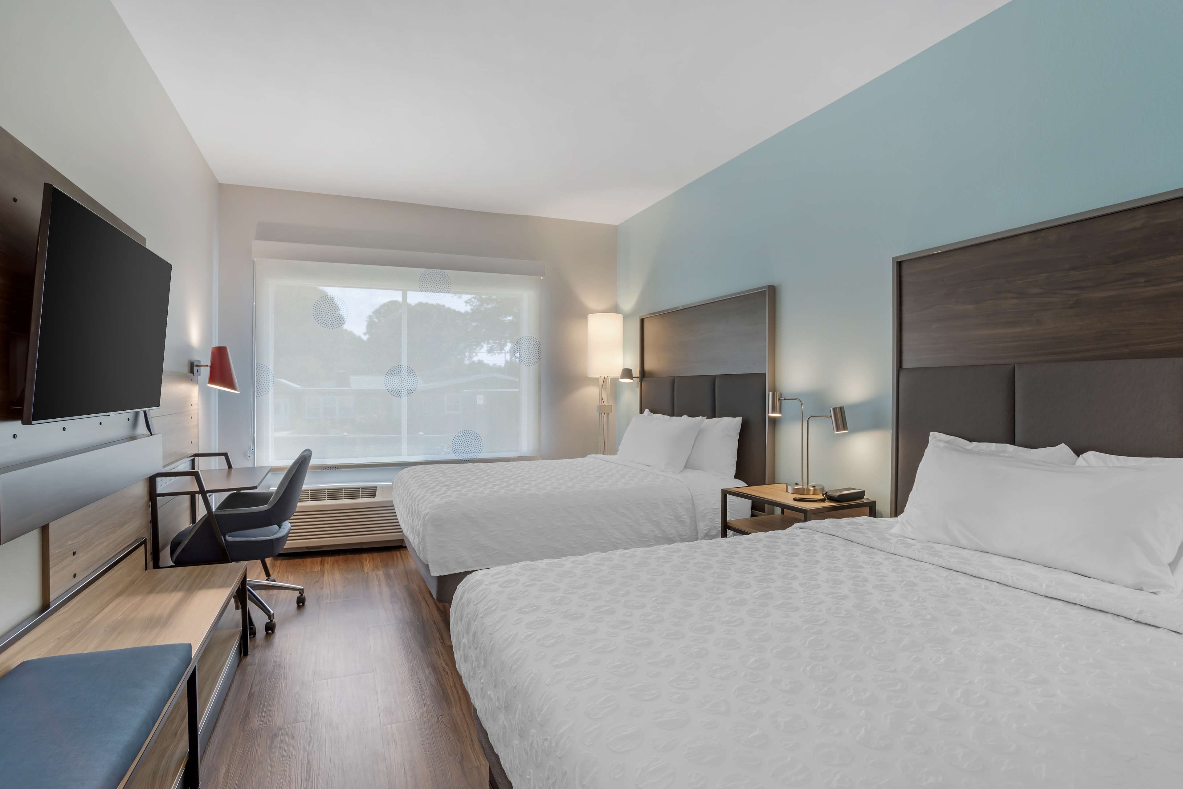 Habitació d'hotel amb dos llits, escriptori amb cadira, televisió i finestra amb vista a arbres.