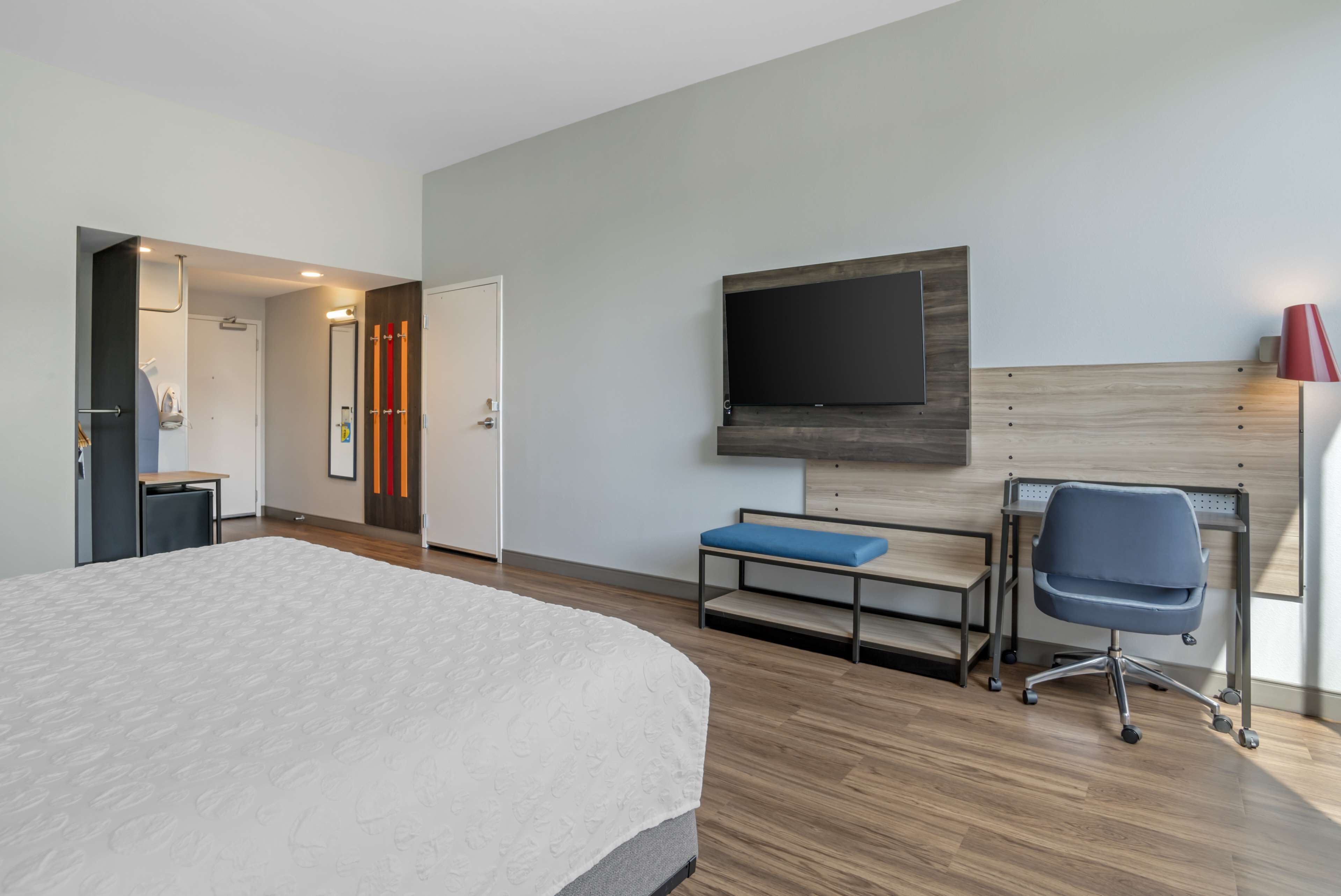 A hotel room with a large bed, a desk, a chair, a TV mounted on the wall, and a bench.