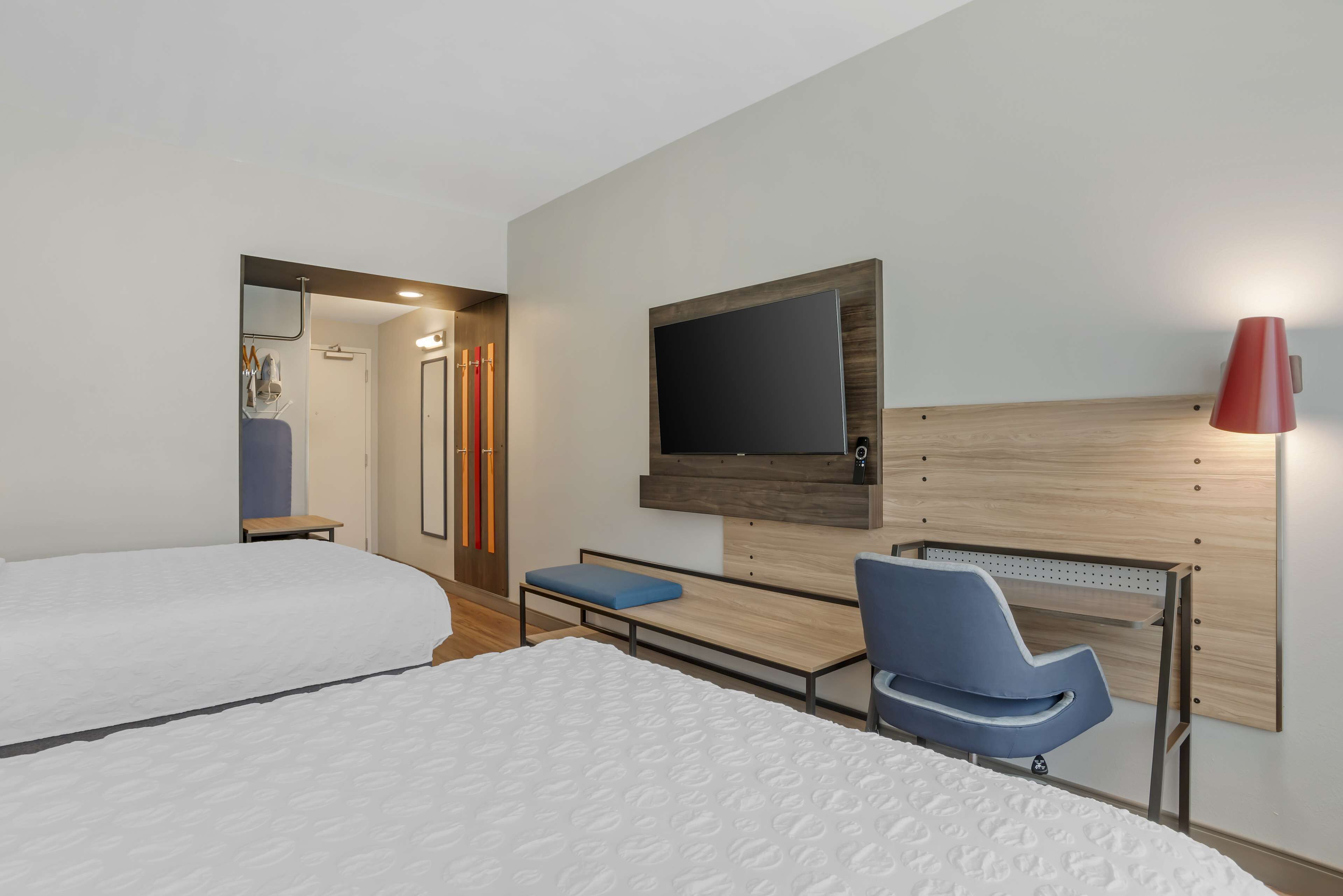 A modern hotel room with two beds, a TV mounted on the wall, a red lamp, and a wooden headboard.