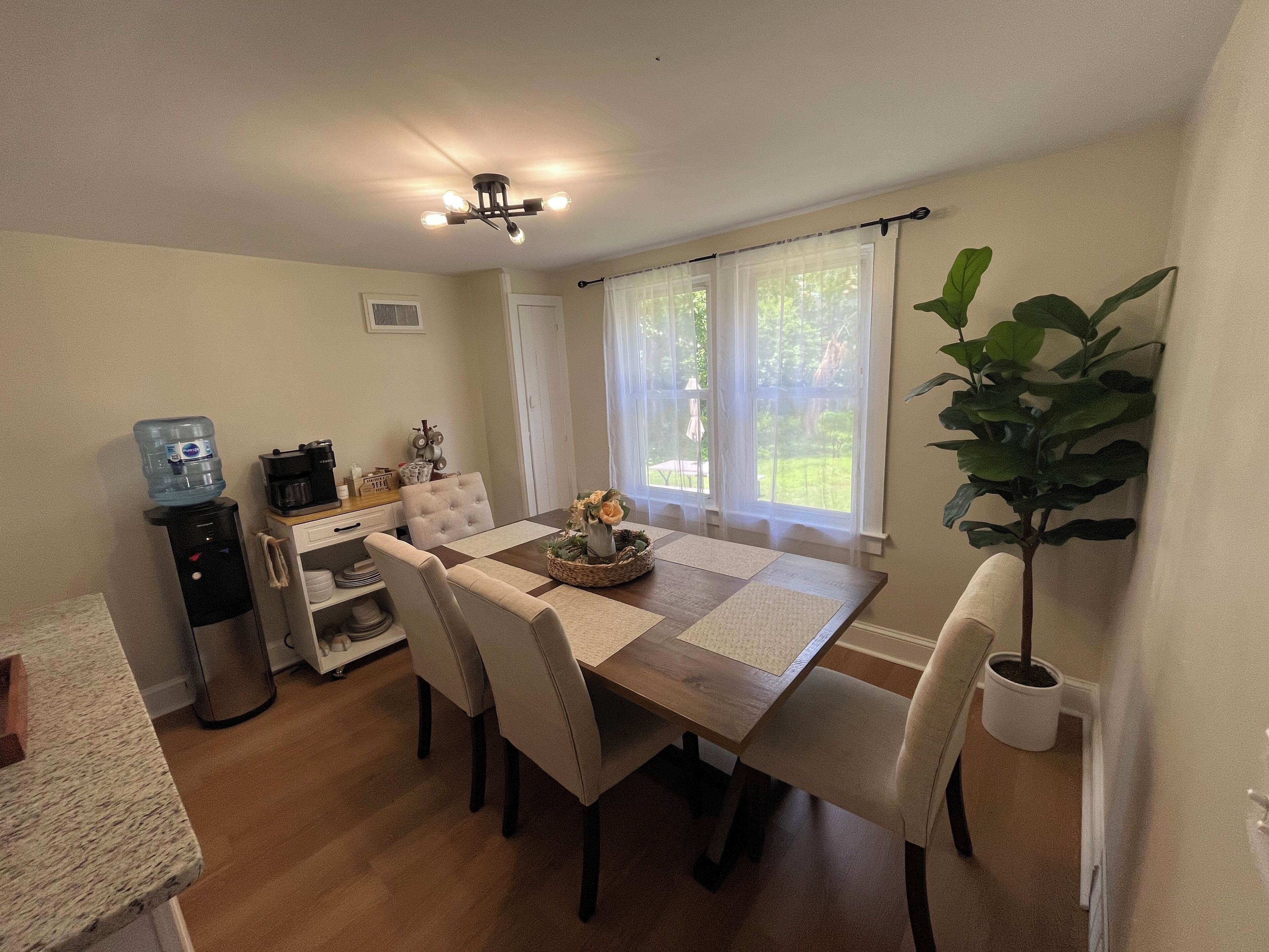 A dining area with a wooden table, four chairs, a water dispenser, a coffee maker, and a potted plant.