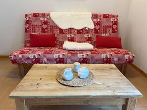 A wooden table with a woven placemat, two white ceramic cups, and a small white pitcher. Behind the table, there is a red patterned sofa with a white throw blanket and two red cushions.