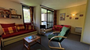 Un salon chaleureux avec un canapé, un fauteuil vert, une table basse en bois et un coin repas avec une table et des chaises rouges.