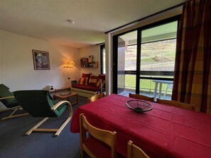 A room with a dining table, chairs, a sofa, a green chair, a small table with a lamp, and a window with a view of the outdoors.