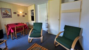 Une pièce comprenant une table à manger, deux chaises vertes rembourrées, une table basse en bois et une bibliothèque.