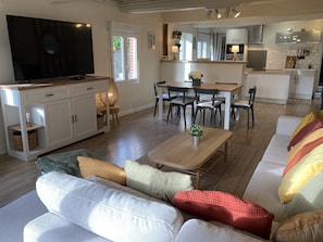 A modern living room with a flat-screen TV, a dining area with a wooden table and chairs, and a kitchen with white cabinets and a built-in oven.