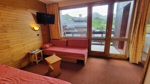Una habitación con pared de madera, un sofá, una mesa pequeña, un televisor y una ventana con vistas a edificios y montañas.