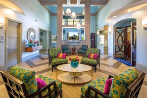 A spacious lobby with a central round table surrounded by patterned armchairs, a reception desk, and a large mirror.