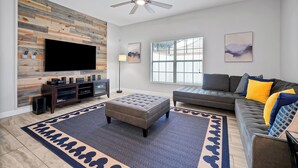 A modern living room with a flat-screen TV, wooden wall paneling, a ceiling fan, a large window, and a gray sofa with colorful cushions. - Make your own Escape to Disney in this fantastic pool townhouse! (Davenport)