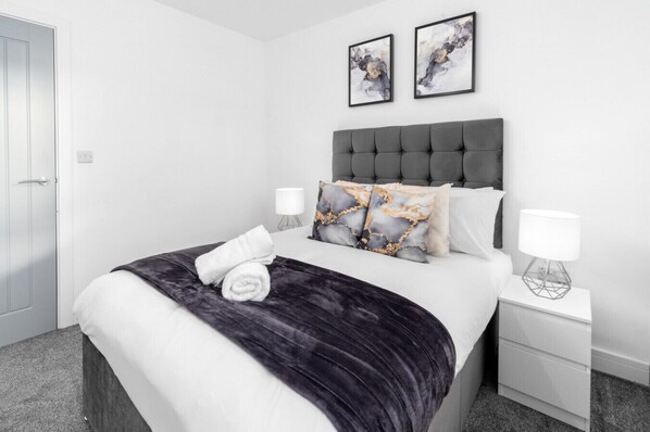 A modern bedroom with a grey upholstered headboard, white bedding, and decorative pillows. There are two bedside tables with lamps and artwork above the bed.