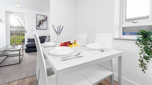 Un'area pranzo moderna con un tavolo bianco apparecchiato per quattro, inclusi piatti, bicchieri e posate. Sullo sfondo, si notano un divano e un tavolino.
