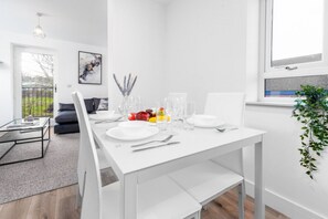 Ein moderner Essbereich mit einem weißen Tisch, gedeckt für vier Personen, einschließlich Teller, Gläser und Besteck. Im Hintergrund sind eine Couch und ein Couchtisch zu sehen.