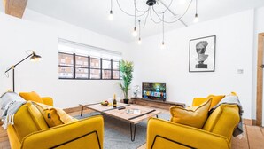 A modern living room with yellow chairs, a wooden coffee table, a potted plant, and a television. - Newtown Lofts - Luxe Apartment - Parking (Birmingham)