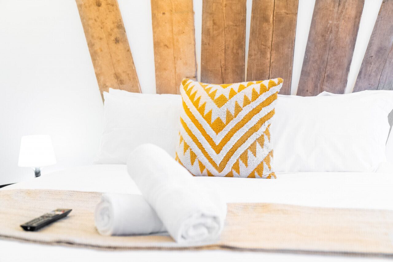 A neatly made bed with white linens, a rolled towel, and a remote control on a wooden headboard.
