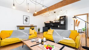 A modern living room with yellow sofas, a wooden coffee table with a fruit platter, and a kitchen area in the background. - Newtown Lofts - Luxe Apartment - Parking (Birmingham)