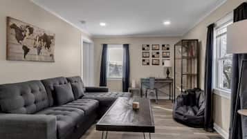 A living room with a sectional sofa, a coffee table, a bookshelf, and a large window with curtains.