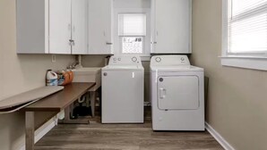 A laundry room with two white washing machines, a countertop with cleaning supplies, and a window with blinds. - Spacious Unit Sleeps 9 + Game Room! 15 Mins from New Bills Stadium (West Seneca)