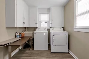 A laundry room with two white washing machines, a countertop with cleaning supplies, and a window with blinds. - 3BR West Seneca Apartment Sleeps 9 w/ Game Room (West Seneca)