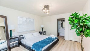 Un dormitorio con cama, mesitas de noche, espejo, ventana con persianas, lámpara en el techo y planta en maceta.