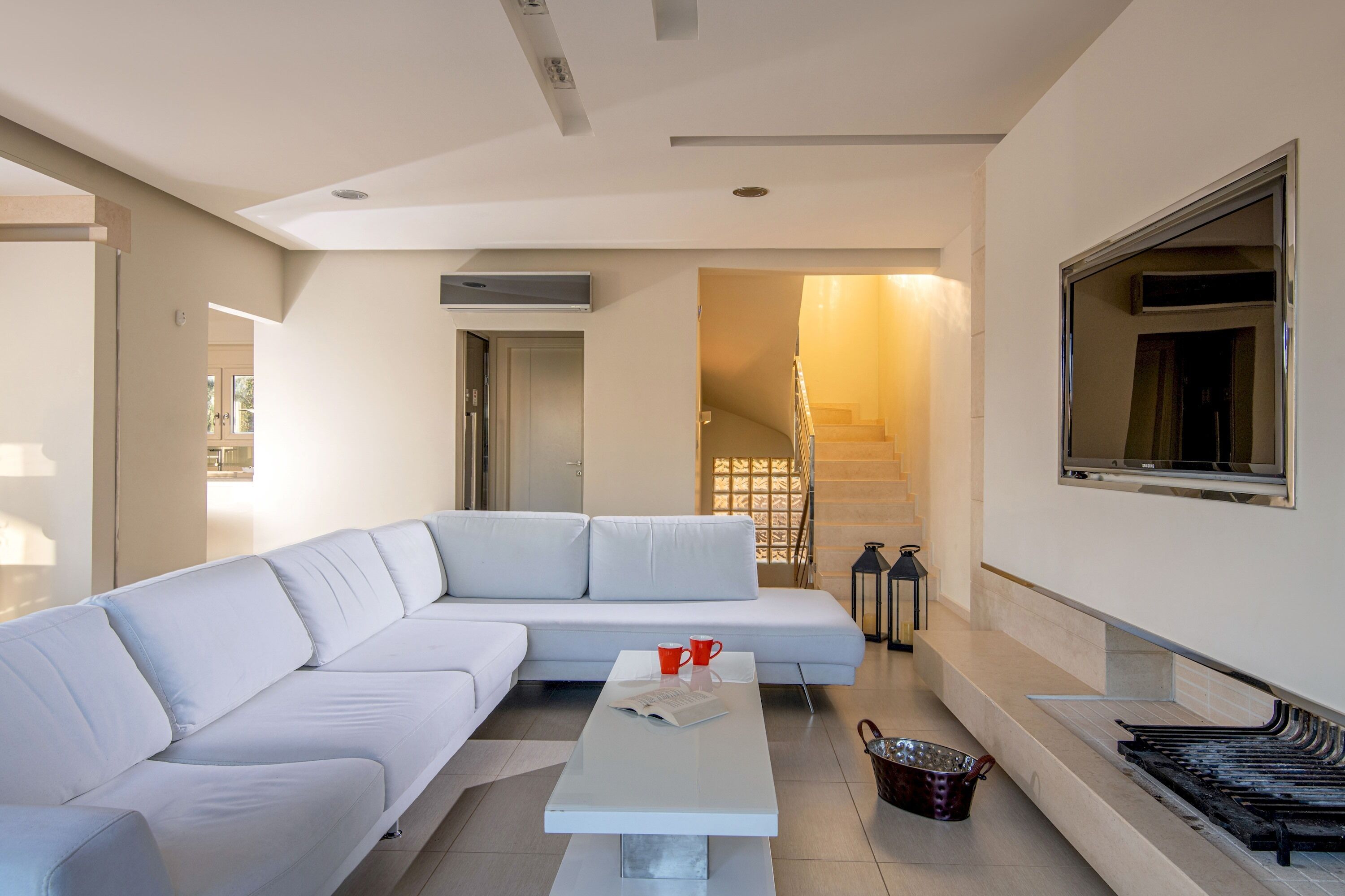Uma sala de estar moderna com um sofá branco modular, uma mesa de centro e uma escada que leva a um andar superior.