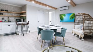 A modern kitchen with white cabinets, a dining area with a round table and teal chairs, and a cozy reading nook with a wicker chair and a television displaying a tropical scene.