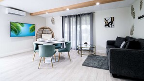 A modern living room with a dining area, featuring a round table, teal chairs, a black sofa, and a flat-screen TV on the wall.