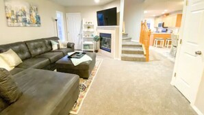 A living room with a sofa, coffee table, and a TV. There is a staircase leading to a kitchen area. - Boise Avenue Getaway • 3BR/3BA • Yard • Parking for 2 • Near BSU & Downtown • Spacious (Boise)