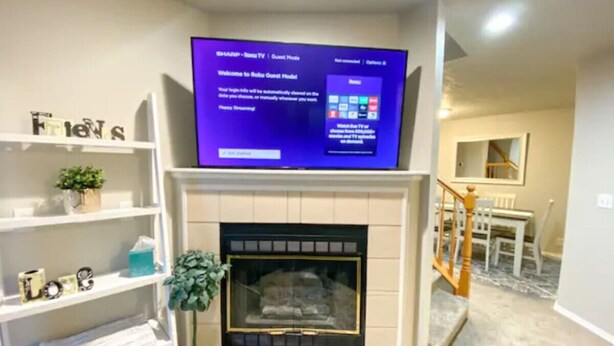 A living room with a fireplace, a mounted flat-screen TV displaying a guest mode welcome screen, and a staircase leading to another room. - Boise Avenue Getaway • 3BR/3BA • Yard • Parking for 2 • Near BSU & Downtown • Spacious (Boise)