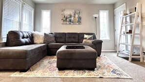 A living room with a sectional sofa, an ottoman, and a floor rug. There is a ladder leaning against the wall and a painting on the wall. - Boise Avenue Getaway • 3BR/3BA • Yard • Parking for 2 • Near BSU & Downtown • Spacious (Boise)