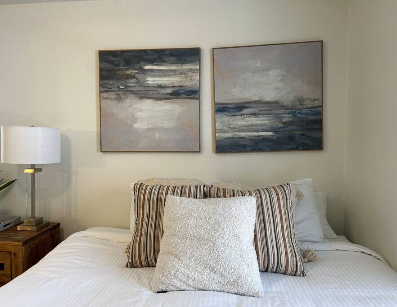 Un dormitorio con una cama, dos cuadros en la pared, una lámpara en una mesita de noche y almohadas a rayas.
