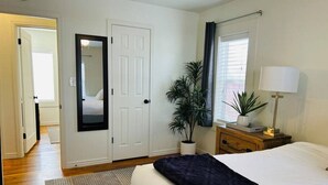 Ein Schlafzimmer mit Bett, Nachttisch, Lampe, Topfpflanze, einer Tür und einem Fenster mit Vorhängen.