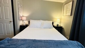 A neatly made bed with white linens and a blue blanket, a nightstand with a lamp, and a framed picture on the wall. - The Red Bench Cottage | Historic Charm • Fenced Yard • Walk to Hyde Park • Central Boise (Boise)