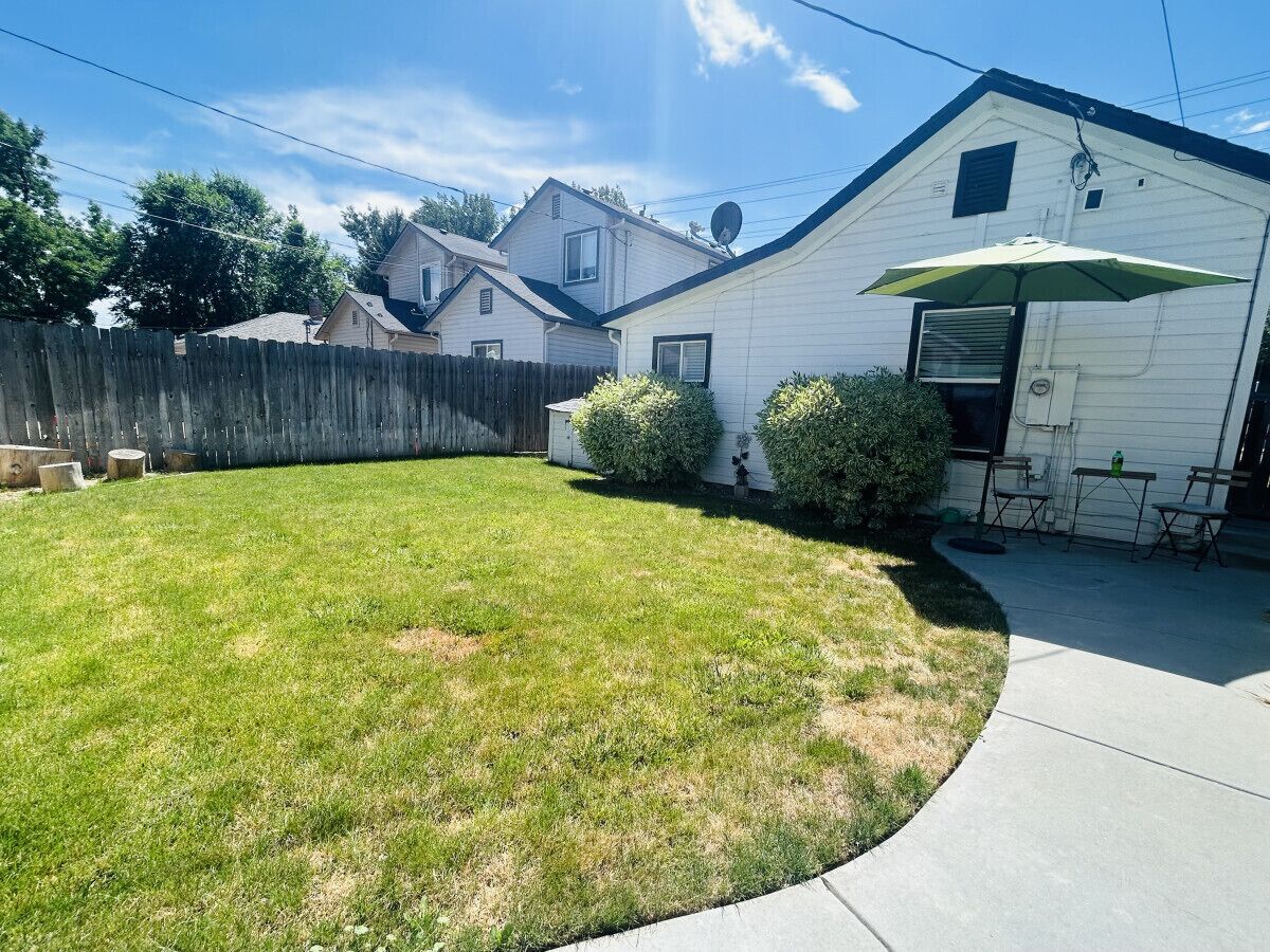 A residential backyard with a lawn, a wooden fence, and a white house with a patio and outdoor furniture.